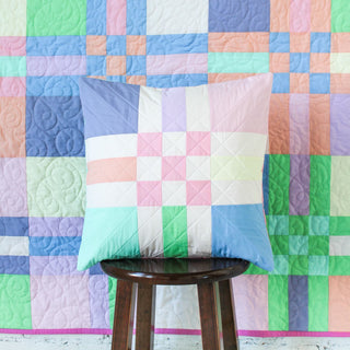 Square patchwork pillow with pastel colors featuring quilted patterns, sitting on a wooden stool in front of a matching pastel patchwork quilt, showcasing To The Nines Quilt Pattern (Download) by Patternly Collective.