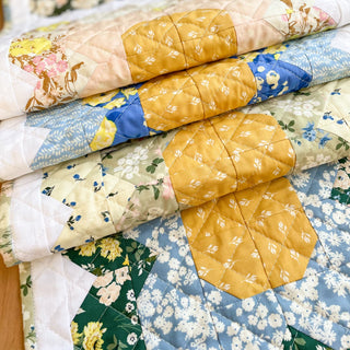 Close-up of folded quilted table runners in floral and geometric prints featuring warm yellow, blue, and green tones, showcasing Trula's Blooms Table Runner Quilt Pattern (Download) by Heart Lake Quilts.
