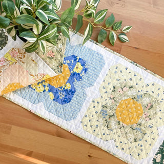 Folded quilt runner with floral patchwork blocks in pastel blue, yellow, and cream tones, styled with green houseplant on a wooden surface by Heart Lake Quilts, promoting Trula's Blooms Table Runner Quilt Pattern (Download).