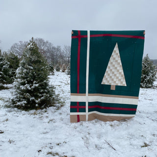 A green quilt featuring a geometric pine tree design held outdoors in a snowy field with evergreen trees, showcasing the Urban Pine Quilt Pattern (Download) by Sew & Sew Quilting Co.