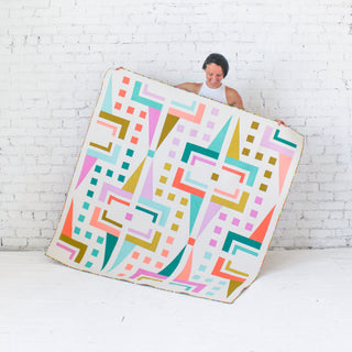 Person holding a colorful geometric-patterned quilt against a white brick wall.