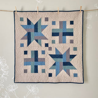 Wink Quilt Pattern (Download) by The Dandy Patch featuring a modern geometric quilt with blue and gray star and cross blocks on a beige background hanging on a wall.
