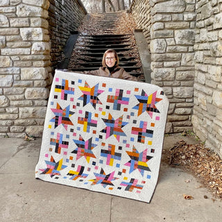 Person holding a colorful star and cross patchwork quilt made with the Wink Quilt Pattern (Download) by The Dandy Patch, displayed outdoors near stone steps.