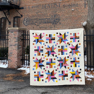 Colorful quilt with star and cross block patterns made from vibrant geometric shapes hanging outdoors against a brick wall, showcasing the Wink Quilt Pattern (Download) by The Dandy Patch.