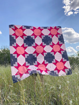 Quilt with pink and blue geometric pattern on a grassy field with blue sky and clouds.