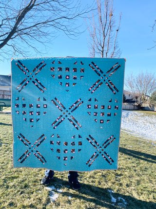 Bright blue quilt with a pattern of hearts and X shapes made from floral fabric, displayed outdoors on a sunny day, showcasing the X My Heart Quilt Pattern (Download) by Sweet Potato Quilts.