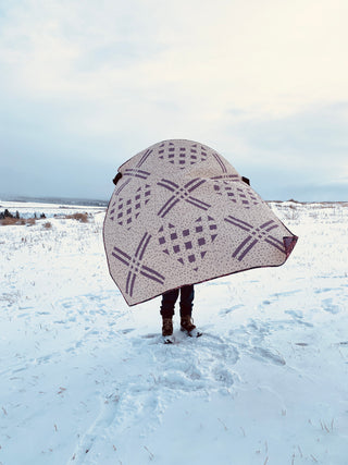 Person holding a round quilt with purple heart and X patterns outdoors on snow, showcasing the X My Heart Quilt Pattern (Download) by Sweet Potato Quilts.
