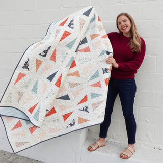 Jubilant Quilt Pattern (Download) by Homemade Emily Jane featuring a modern quilt with colorful triangular shapes and playful negative space displayed by a smiling woman