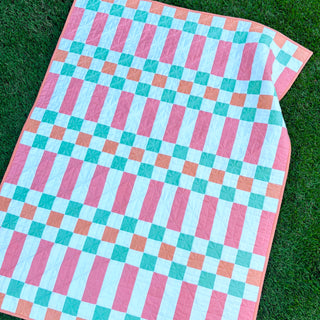 Linear Quilt Pattern (Download) by Homemade Emily Jane featuring a modern geometric quilt with pink blue and teal stripes arranged in a crisp grid on green grass