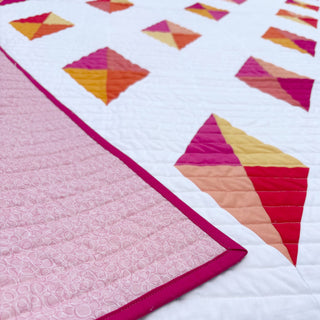 Luminary Quilt Pattern (Download) by Homemade Emily Jane showing a white quilt with colorful geometric triangles and a pink patterned backing